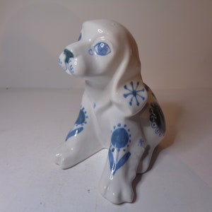 Può includere: Una statuetta di ceramica bianca di un cucciolo seduto con disegni floreali blu e bianchi. Il cucciolo ha gli occhi blu e il naso nero.