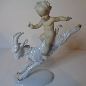 May include: A white porcelain figurine of a cherub riding a leaping goat. The cherub is nude and has a mischievous expression. The goat has gold accents on its hooves.