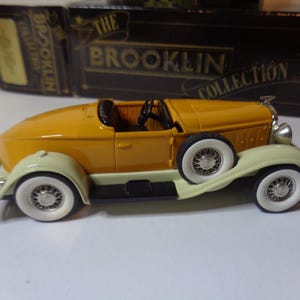 Könnte beinhalten: Ein gelbes Vintage-Automodell aus den 1930er Jahren mit weißen Akzenten. Das Auto ist ein Cabriolet mit schwarzem Innenraum. Das Modell steht auf einer schwarzen Oberfläche. Das Logo der Brooklyn Collection ist im Hintergrund sichtbar.