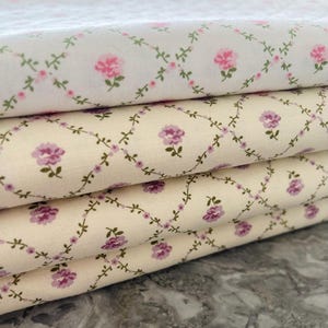 May include: A stack of floral fabric featuring a repeating pattern of pink and purple roses, linked by a green vine design. The fabric has a white and cream background.