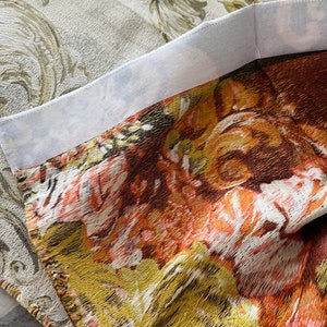 May include: Close-up of a fabric with a floral pattern in shades of orange, yellow, and brown. The fabric has a textured surface and a white border.