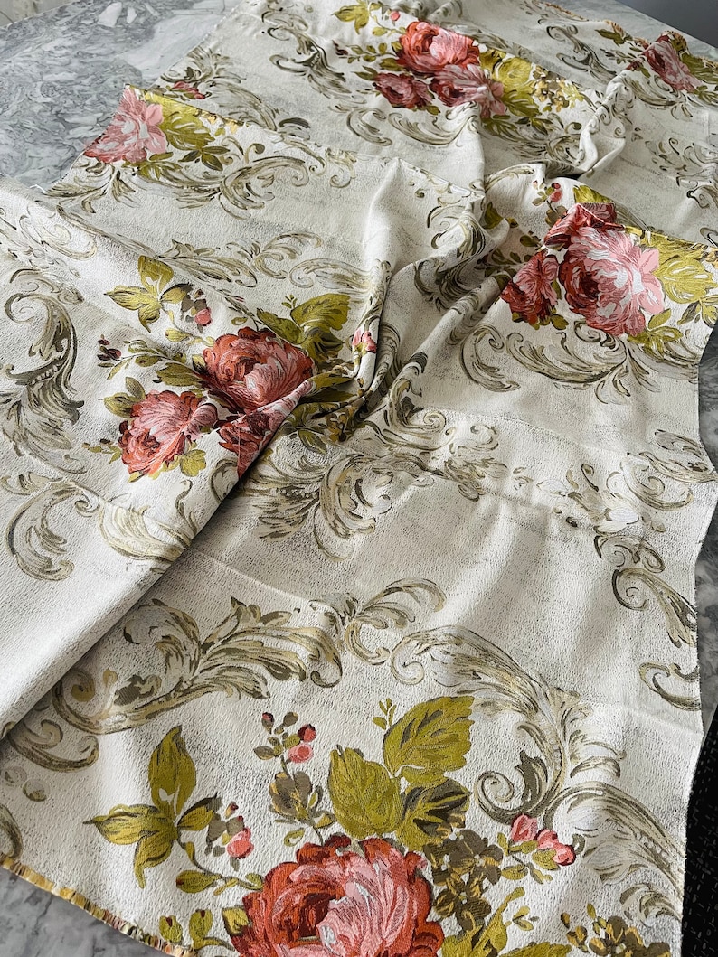 May include: A white fabric with a floral pattern. The fabric features a repeating pattern of pink and red roses with green leaves and stems. The background is a light beige color with a subtle texture.