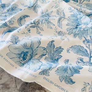 May include: White fabric with a blue floral pattern. The fabric has a frayed edge and the text "English Garden Floral" is printed on the bottom edge. The text "English Heritage by Designers Guild" is printed on the top edge.