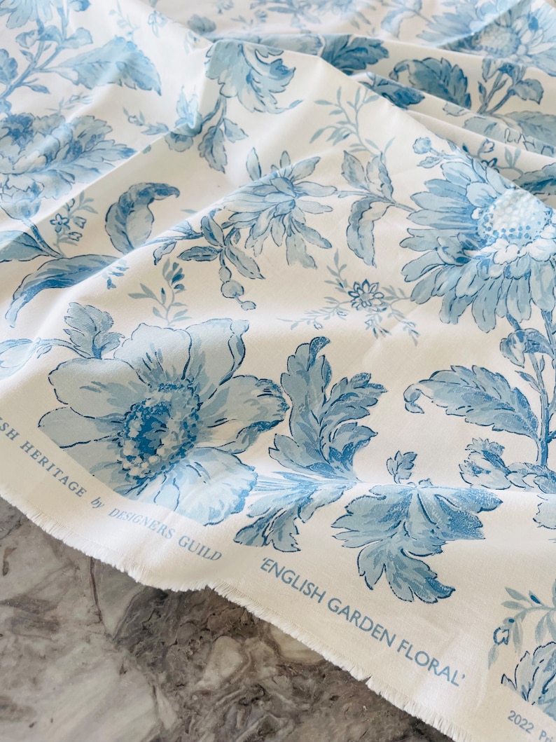 May include: White fabric with a blue floral pattern. The fabric has a frayed edge and the text "English Garden Floral" is printed on the bottom edge. The text "English Heritage by Designers Guild" is printed on the top edge.