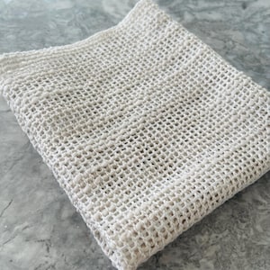 May include: An off-white, square mesh produce bag. The bag is made of a loosely woven material, with a grid-like pattern. The bag is folded and appears to be empty. The background is a light grey, marble-like surface.
