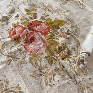 May include: A close-up of a white fabric with a floral pattern. The fabric features a repeating pattern of gold and brown leaves and pink roses. The fabric has a textured surface.