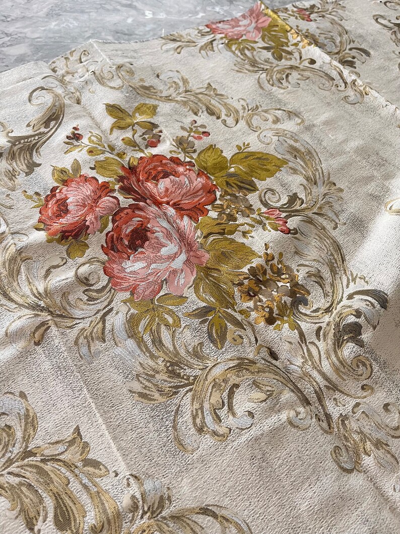 May include: A close-up of a white fabric with a floral pattern. The fabric features a repeating pattern of gold and brown leaves and pink roses. The fabric has a textured surface.