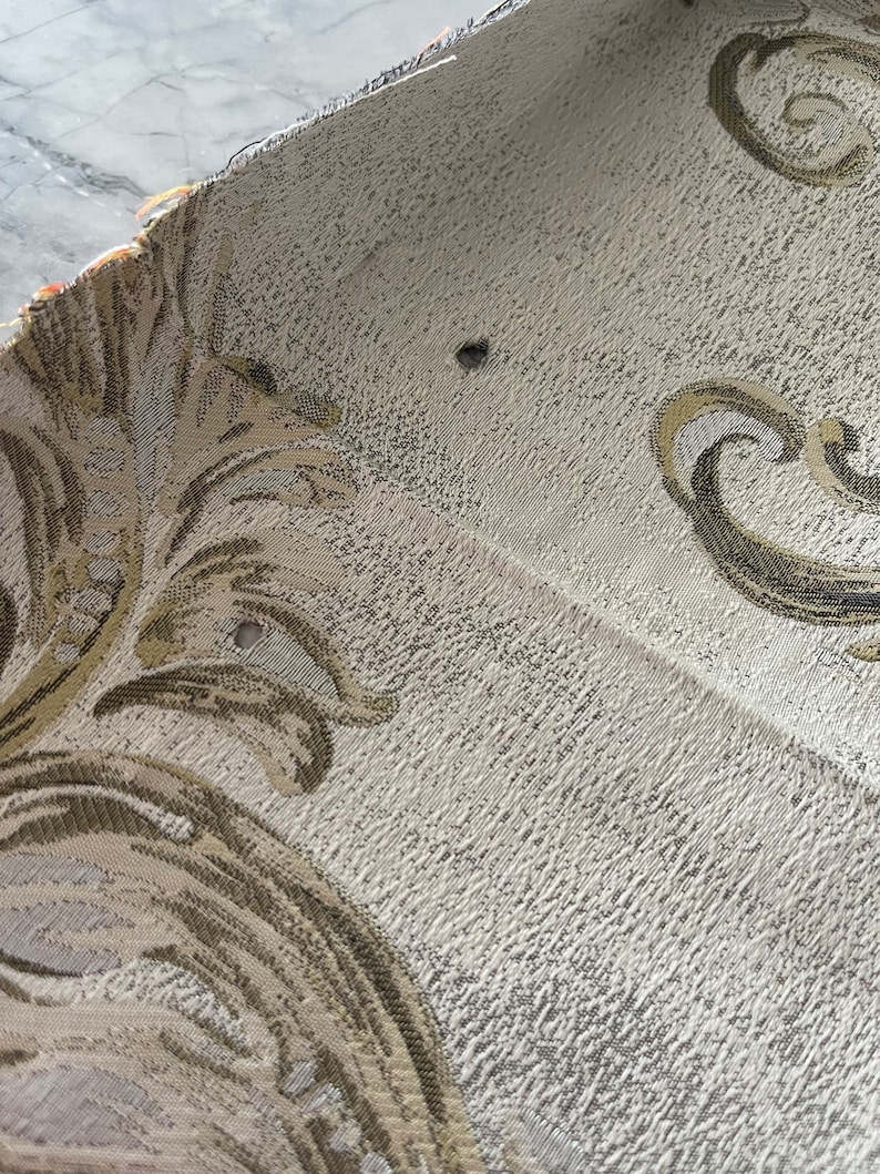 May include: Close-up of a beige fabric with a gold and brown floral pattern. The fabric has a textured surface and appears to be woven.