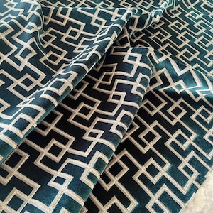 Op de afbeelding: Close-up van een gevouwen teal stof met een geometrisch patroon. Het ontwerp heeft een herhalend patroon van witte, onderling verbonden, hoekige vormen. De stof heeft een zachte, fluweelachtige textuur en de plooien creëren schaduwen.