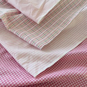 May include: A close-up shot of layered pink and white fabrics. The fabrics display a variety of patterns, including stripes, gingham, and a small plaid design. The fabrics are folded and layered, highlighting the different textures and patterns.