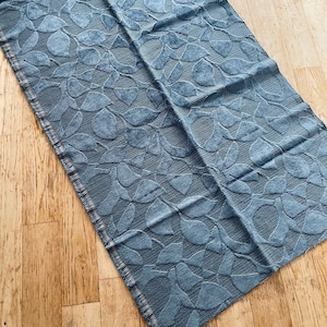 May include: A rectangular blue fabric runner featuring a raised, textured leaf pattern across the entire surface. The material displays a consistent denim-like blue hue with a woven structure and finished edges along the sides of the textile piece.
