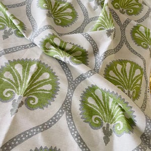 May include: Cream-coloured fabric featuring a repeating pattern of green and grey floral motifs. The floral elements are outlined in grey and linked by a dotted grey border. The fabric is folded, displaying its texture and pattern.