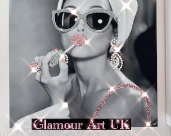 May include: Black and white photo of a woman wearing sunglasses and a towel on her head. She is holding a lipstick and applying it to her lips. The photo is covered in glitter and the text "Glamour Art UK" is at the bottom.