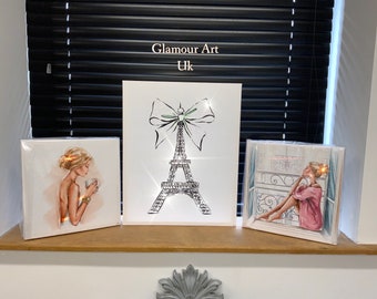 May include: Three framed canvas prints with a Parisian theme. The centre print features a black and white illustration of the Eiffel Tower with a green and white bow. The left print shows a woman in a pink dress holding a cup of coffee. The right print shows a woman in a pink robe sitting by a window.