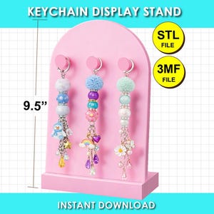 Op de afbeelding: Een roze boogvormige standaard voor sleutelhangers van 24,13 cm hoog, met drie haken waaraan kralen sleutelhangers met pompons en bedels hangen. De tekst luidt KEYCHAIN DISPLAY STAND, STL FILE, 3MF FILE en INSTANT DOWNLOAD.