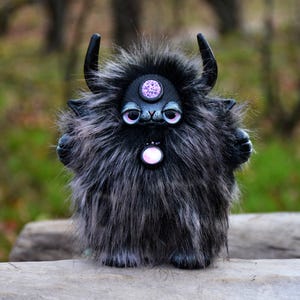 May include: A black and gray furry creature with horns and a pink gem on its forehead. The creature has large, round, pink eyes and a pink gem on its chest.