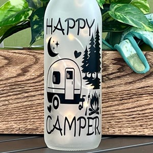May include: Frosted glass bottle with the words "HAPPY CAMPER" and a black graphic of a camper, trees, moon, and campfire. The bottle is lit with small lights, creating a warm glow. The bottle is a decorative item.
