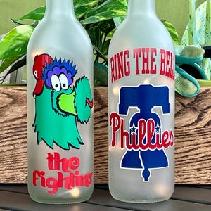 May include: Two frosted glass bottles with Phillies baseball team logos and text. One bottle features the Phillie Phanatic mascot and the words "the Fightins". The other bottle has "Ring the Bell" and the Phillies logo.