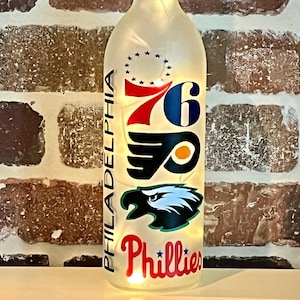 May include: A clear glass bottle with string lights inside, featuring logos for Philadelphia sports teams. The bottle has the text "PHILADELPHIA" vertically and logos for the 76ers, Flyers, and Phillies.