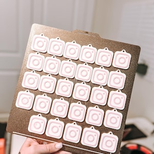 May include: A collection of pink and white Instagram camera-shaped charms arranged on a brown rectangular base. Each charm has a small hole at the top. The charms are held by a person.