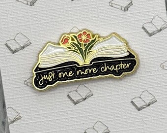 May include: A gold-colored enamel pin in the shape of an open book with the text "just one more chapter." The pin features white pages, black borders, and a floral design with red and yellow flowers. The pin is on a patterned background.