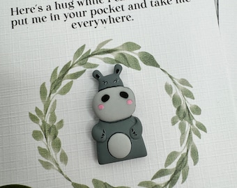 May include: A gray hippo-shaped charm with pink cheeks and a white belly button is centered on a card. The card has the text "Pocket Hippo" and a message about carrying it everywhere. A green wreath surrounds the hippo.