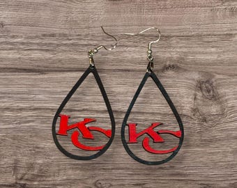 May include: A pair of teardrop-shaped earrings featuring a black outline and a red KC logo centered within each frame. The earrings are suspended from silver-toned metal fishhook wires, creating a distinct design that highlights the bold red lettering.