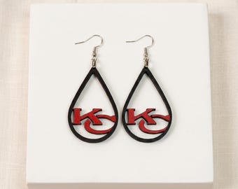 May include: A pair of teardrop-shaped earrings featuring a black outline and a red KC logo in the center. The earrings are suspended from silver-toned metal hooks. The design is symmetrical, with the letters K and C overlapping in a bold font.