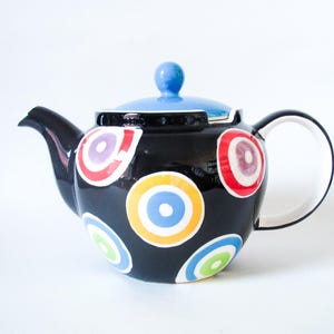 May include: A black ceramic teapot with a blue lid and handle. The teapot is decorated with colorful, concentric circle patterns in red, yellow, purple, and green. The spout and handle are black and white.