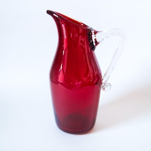 May include: A ruby red glass pitcher with a clear glass handle. The pitcher has a curved spout and a rounded body. The handle is attached with a small glass bead. The pitcher is likely used for serving beverages.