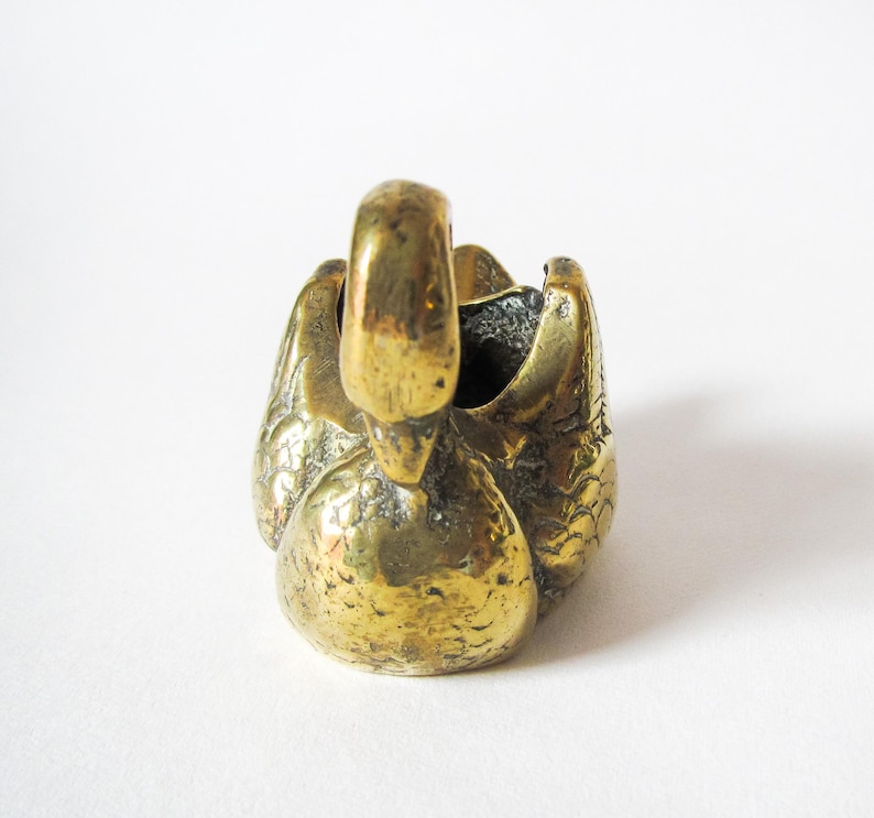 May include: A brass swan figurine with a detailed, textured surface. The swan is depicted with its head raised and wings folded, creating a small, open bowl shape. The brass has a warm, golden hue, and the overall design is compact and decorative.