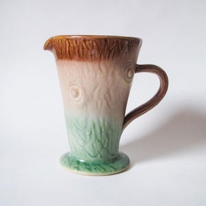 May include: A brown, pink, and green ceramic pitcher with a tree bark design. The pitcher has a wide base and a handle.