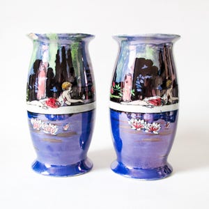 May include: A pair of iridescent ceramic vases featuring a hand-painted scene of a reclining figure in a landscape with dark trees above a band of white water lilies floating on a deep blue pond. The vases have a flared rim and base.