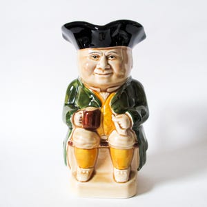 May include: A ceramic Toby jug figurine of a seated man in a tricorn hat, green coat, and yellow vest. The jug holds a brown mug and has a smiling face. The base is rectangular.