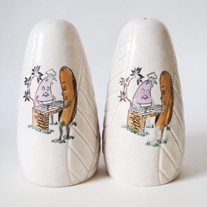 May include: A pair of ceramic salt and pepper shakers. Each shaker is off-white with a leaf-like pattern. The shakers feature a cartoon illustration of a hot dog and a purple onion grilling. The hot dog and onion are standing at a brick grill.