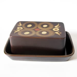 May include: A brown ceramic butter dish with a rectangular lid featuring a decorative pattern of circles and a red accent. The dish sits in a matching rectangular base, perfect for serving butter.