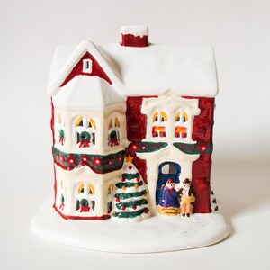 May include: A ceramic Christmas house figurine with a red exterior, white roof, and snow-covered base. The house features windows, a Christmas tree, and a door with figures. Festive garland and a chimney complete the holiday scene.