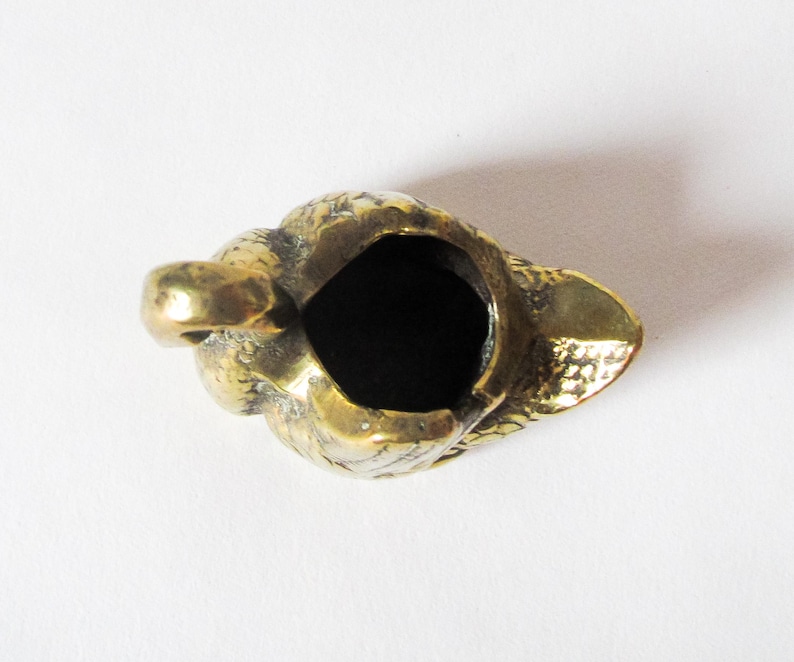May include: A small, antique brass ashtray with a textured surface. The ashtray is shaped like a bird's head, with a deep, dark interior. The bird's beak forms the front of the ashtray, and the head is detailed with feathers.