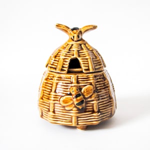 May include: A glazed ceramic honey pot shaped like a traditional woven beehive. The golden-brown container features a textured basket-weave pattern, a removable lid with a small handle, and a raised relief of a bumblebee on the front surface.