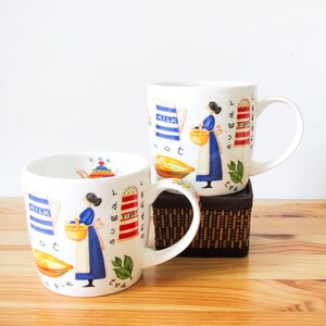 May include: Two white ceramic mugs featuring colorful illustrations of a person in a blue dress holding a yellow basket, a blue and white striped milk pitcher, a red and white striped sugar shaker, a pie, and the words milk, sugar, tea, and pie.