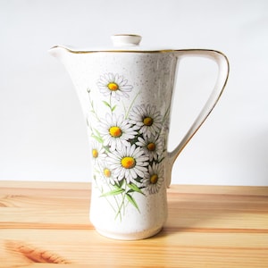 May include: A white ceramic pitcher with a lid, featuring a daisy floral design. The pitcher has a curved handle and a gold trim. The daisies have white petals and yellow centers, with green leaves and stems.