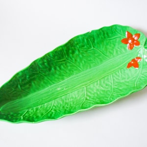 May include: A ceramic serving dish shaped like a large green leaf with detailed veining. Two small red flowers are painted on the upper right side of the leaf. The dish is on a white background.