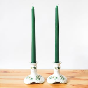 May include: Two tall, slender dark green taper candles are held upright in matching white ceramic candle holders. Each holder features a wide, scalloped base decorated with painted green leaves and small white flowers, providing a stable foundation for the candles.