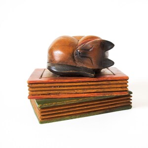 May include: A hand-carved wooden figurine of a sleeping cat resting on top of two stacked wooden boxes. The cat is finished in a warm brown tone with darker accents on the ears and tail. The boxes feature orange and green painted edges.