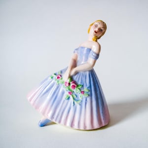 May include: A porcelain figurine of a woman in a flowing blue and pink dress, holding a bouquet of pink roses. The figurine has blonde hair and is posed in a graceful stance. A decorative item.