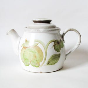 May include: White ceramic teapot with a lid and handle. The teapot features a green and yellow floral design. The spout and handle are a light beige colour. The lid is a darker brown colour.