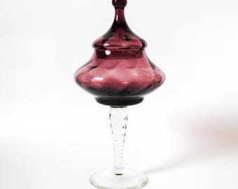 May include: A tall, elegant glass jar with a lid. The jar is a deep, translucent purple, and the stem is clear with a twisted design. The jar has a bulbous body and a rounded lid with a small knob on top. The base is a flat, circular foot.