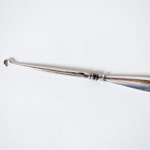 May include: A silver surgical instrument with a curved hook at one end and a rounded handle. The instrument is elongated and tapers towards the hook. The handle has decorative detailing. The object is set against a white background.