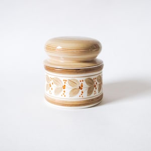 May include: A ceramic jar with a lid. The lid and the top band of the jar are a light brown color with horizontal stripes. The jar has a white base with a brown floral pattern and orange accents.