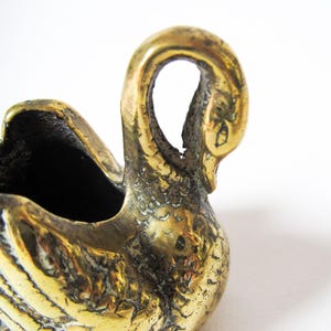 May include: A vintage brass swan figurine with a textured surface. The swan has a curved neck and a hollowed-out body, possibly for holding small items. The brass has a warm, golden hue, and the swan is set against a white background.